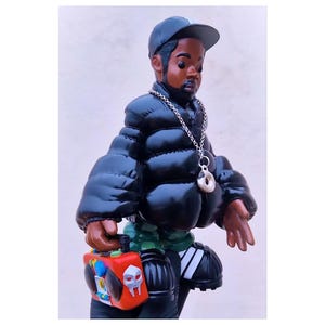 Könnte beinhalten: Eine schwarze und grüne Spielzeugfigur, die eine schwarze Steppjacke, eine schwarze Baseballkappe und eine silberne Kette mit einem Donut-Anhänger trägt. Die Figur hält eine rote und schwarze Boombox.