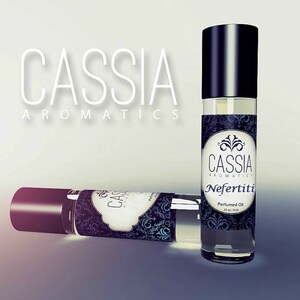 May include: Two black and clear glass roll-on bottles of perfume oil with a black and white label that says "Cassia Aromatics Nefertiti Perfumed Oil 13 oz / 18 ml".