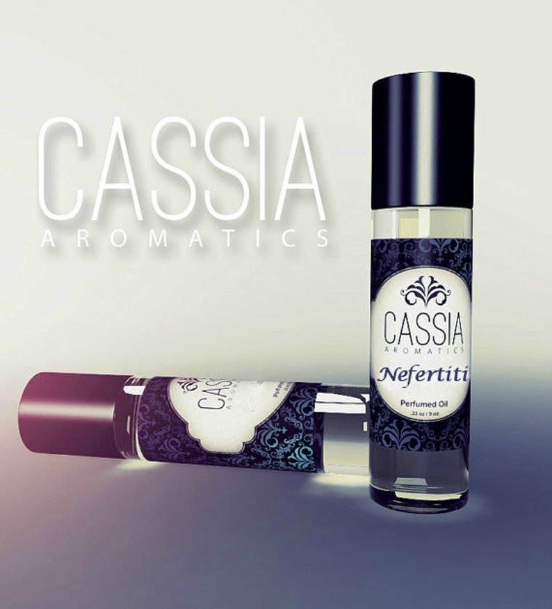 May include: Two black and clear glass roll-on bottles of perfume oil with a black and white label that says "Cassia Aromatics Nefertiti Perfumed Oil 13 oz / 18 ml".