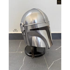 May include: A silver-colored helmet with a black visor, displayed on a black stand. The helmet has a distinctive design with angular features and a smooth, metallic finish. The helmet is likely a prop or collectible.