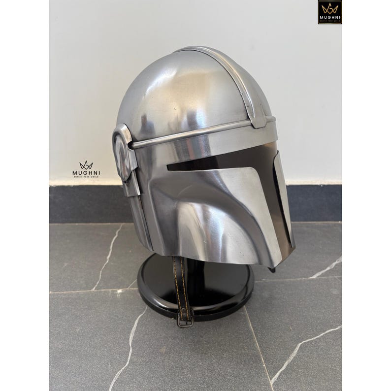 May include: A silver-colored helmet with a black visor, displayed on a black stand. The helmet has a distinctive design with angular features and a smooth, metallic finish. The helmet is likely a prop or collectible.