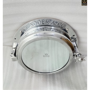May include: A polished silver porthole with the text "CHADDER & CO" engraved on the top. The porthole has a round glass window and a hinged design with a star-shaped handle. The brand logo "MUGHNI" is visible.