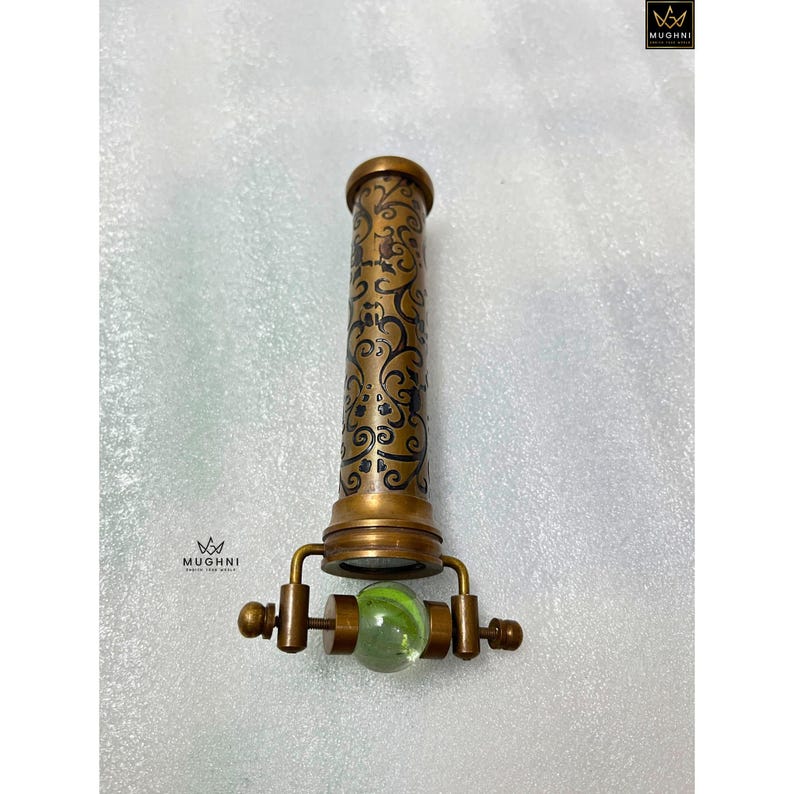 May include: A vintage brass kaleidoscope with an ornate, swirling pattern etched on the barrel. The kaleidoscope features a green marble at the viewing end and a decorative handle. The product is from the brand "Mughni".