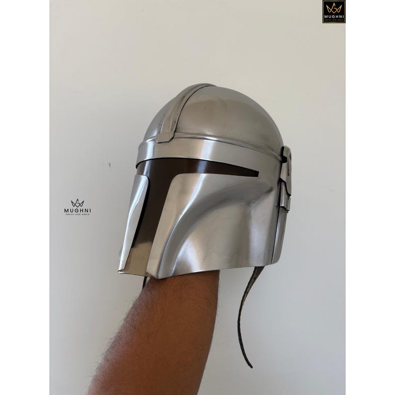 May include: A silver-colored helmet with a dark visor and a sleek, futuristic design. The helmet has a smooth, metallic finish and is likely part of a costume or cosplay item. The helmet is held up by a person's arm.