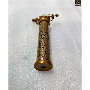 May include: A vintage-style brass kaleidoscope with intricate floral patterns etched on its body. The kaleidoscope features a rotating eyepiece and a small, glowing element at the top. The design includes a small amount of text.