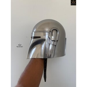 May include: A silver-colored helmet with a visor and a distinctive design. The helmet has a smooth, metallic finish and a strap. The helmet is held up against a white background.