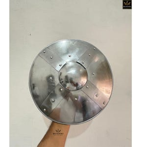May include: A circular silver-coloured metal shield featuring a central domed boss and two intersecting metal bands secured with rivets. The surface has a polished, reflective finish, and the design includes the Mughni logo in the corner.