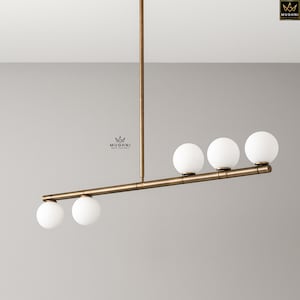 May include: A contemporary pendant light with a bronze finish. It has four spherical, white glass globe shades along a horizontal bar. The light hangs from a single vertical rod, suitable for a modern interior.