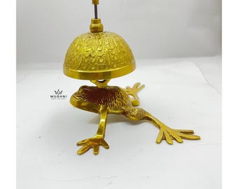 May include: A brass frog-shaped bell with a detailed, textured design. The bell has a dome shape and a small handle on top. The frog's legs are outstretched, and the bell is supported by the frog's back. The bell is a decorative item.