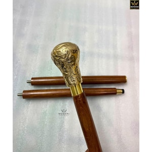 May include: A wooden walking cane with a decorative brass handle. The handle features an ornate floral design. The cane is brown with a brass collar. Two additional sections of the cane are visible.