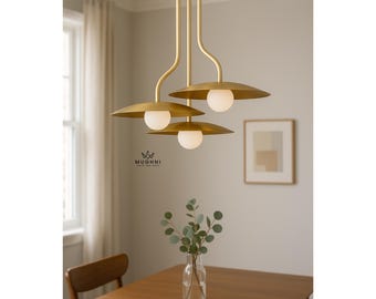 May include: A modern gold-toned pendant light fixture with three lights. Each light has a round, white globe and a wide, saucer-shaped shade. The fixture hangs above a wooden table, with a vase of greenery in the foreground. The brand name "MUGHNI" is visible.