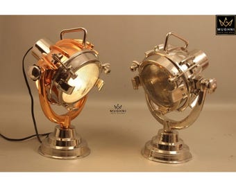 May include: Two industrial-style spotlights. One is copper-colored, the other is silver. Both have a round light, a handle, and a stand. The spotlights are on a neutral background.