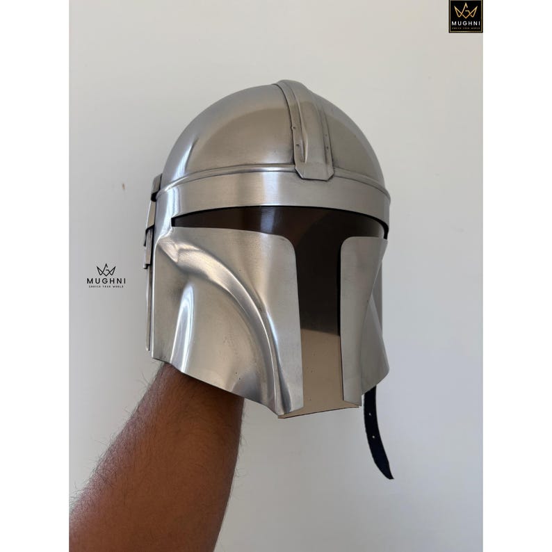 May include: A silver-colored helmet with a distinctive T-shaped visor. The helmet has a smooth, metallic finish and a curved design, with a dark interior visible through the visor. The helmet is held up by a person's arm.
