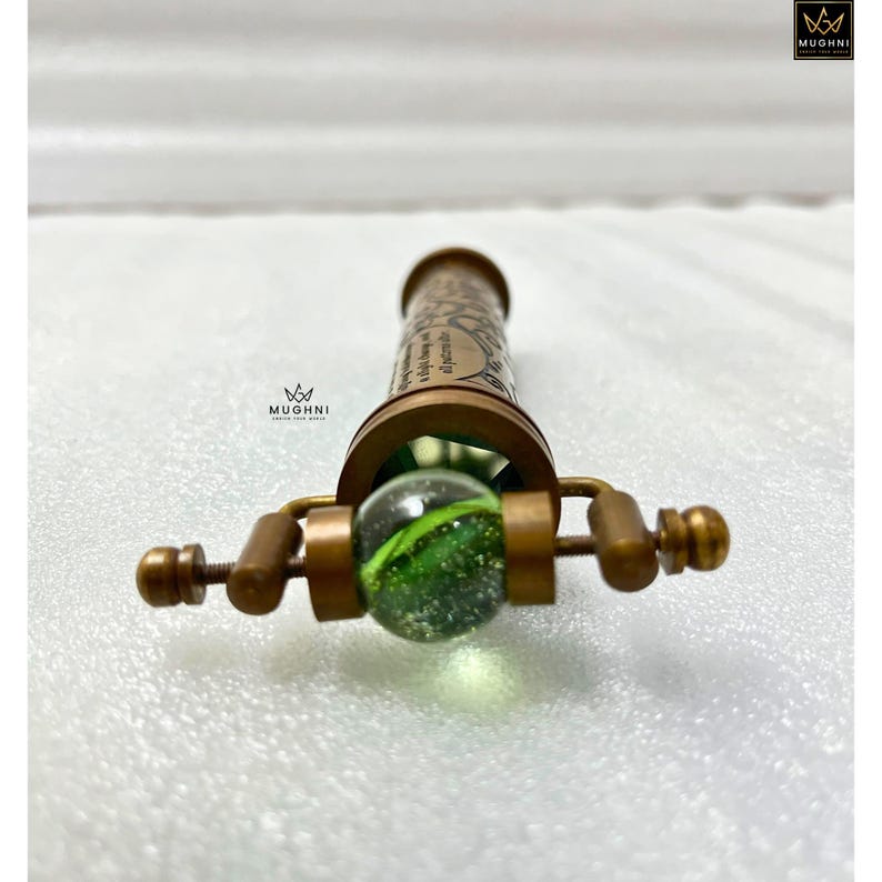 May include: A vintage brass kaleidoscope with a green glass marble at the end. The kaleidoscope has decorative etchings and text. The brass has a warm, antique finish. The product is from the brand Mughni.