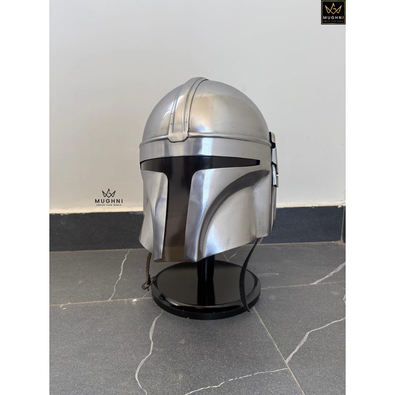 May include: A silver-colored Mandalorian helmet with a black visor, displayed on a black stand. The helmet has a smooth, metallic finish and is designed with distinct angular features. The helmet is a replica of the one from the popular Star Wars series.