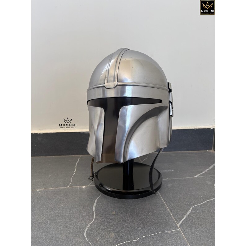 May include: A silver-colored Mandalorian helmet, displayed on a black stand. The helmet has a sleek, metallic finish with a distinctive visor and cheek plates. The helmet is a replica of the one worn by the character in the Star Wars series.