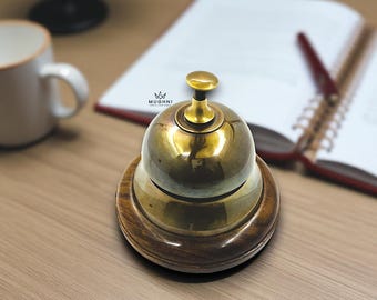 May include: A brass desk bell with a wooden base. The bell has a rounded top and a small knob. The wooden base is circular and has a dark brown finish. The bell is sitting on a wooden desk.