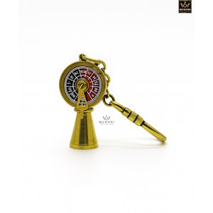 May include: A brass-colored keychain featuring a miniature ship's wheel design. The wheel has a red and black dial with directional markings. The keychain includes a chain and a handle-like extension. The base is a cone shape.