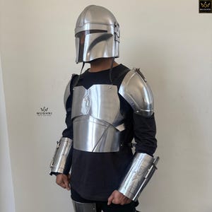 May include: A silver-colored Mandalorian armor set, including a helmet, chest plate, shoulder pads, and arm guards. The armor is made of metal and features intricate detailing. The person is wearing a black long-sleeve shirt underneath the armor.