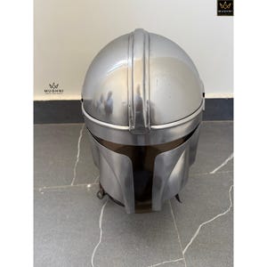 May include: A silver-colored helmet with a visor. The helmet has a rounded top with a central ridge and a smooth, metallic finish. The visor is a darker shade, and the helmet appears to be made of metal. The helmet is a prop or costume piece.