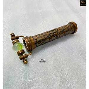 May include: A vintage brass kaleidoscope with intricate floral patterns and a quote: "Nothing remains the same, a slight change, and all patterns alter." The kaleidoscope features a green and clear glass marble at one end.