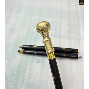 May include: A black walking cane with a gold-colored handle and accents. The handle has an ornate floral design. The cane is in three sections, with a screw-on tip. The logo "MUGHNI" is in the upper right corner.