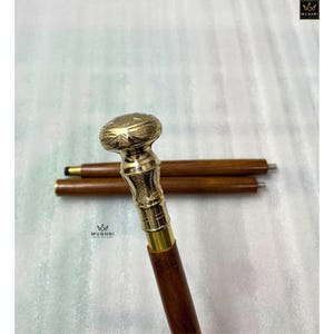 May include: A decorative walking cane with a brass handle and a dark brown wooden shaft. The handle has an engraved floral design. Two additional cane sections are visible in the background.