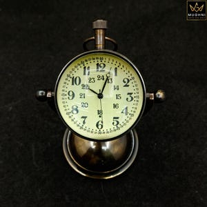 May include: A vintage brass clock with a cream face and black numerals. The clock has a round face with a detailed minute track and a small handle. It sits on a brass base. The clock's diameter is approximately 7 cm.