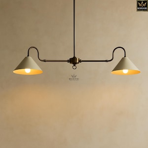 May include: A two-light pendant lamp with a vintage aesthetic. The lamp features two off-white cone-shaped shades with gold interiors, suspended from a dark metal frame. The lamp is lit, casting a warm glow. The brand name "MUGHNI" is visible.