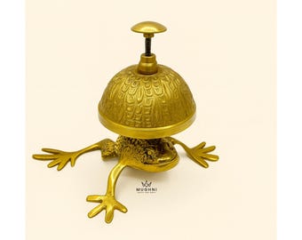 May include: A brass frog-shaped bell with a domed top and a small handle. The bell has a detailed, textured surface and outstretched legs. The frog bell is a decorative item.