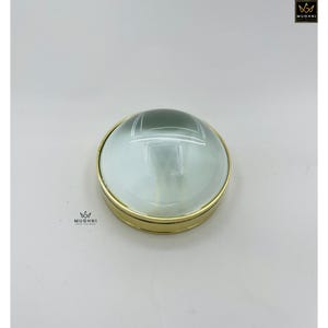 May include: A round magnifying glass with a gold-colored metal frame. The glass is clear with a slight green tint. The magnifying glass is sitting on a white surface. The brand name "MUGHNI" is printed on the top right and bottom left.