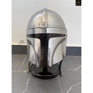 May include: A silver-colored Mandalorian helmet with a black visor and a black stand. The helmet has a smooth, metallic finish and is displayed against a neutral background. The helmet is a replica of the one worn by the character in the Star Wars series.