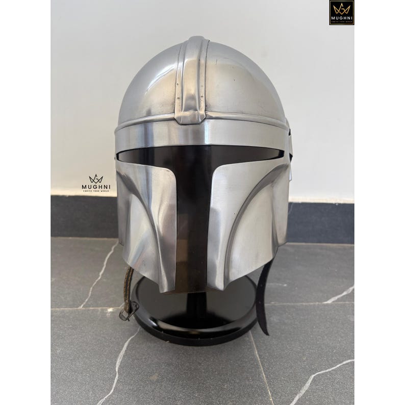 May include: A silver-colored Mandalorian helmet with a black visor and a black stand. The helmet has a smooth, metallic finish and is displayed against a neutral background. The helmet is a replica of the one worn by the character in the Star Wars series.