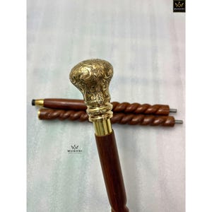 May include: A decorative walking cane with a brass handle and a dark brown wooden shaft. The handle is intricately designed with floral patterns. Two additional cane shafts are visible in the background.