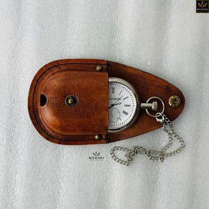 May include: A classic pocket watch with a white face and black numerals, marked "WALTHAM", is displayed in a brown leather case. A silver chain is attached. The case is oval and has brass rivets.