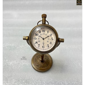 May include: A vintage-style brass clock with a round face and a stand. The clock face has black numerals and hands, and the stand has an ornate design. The clock is approximately 6 inches tall.