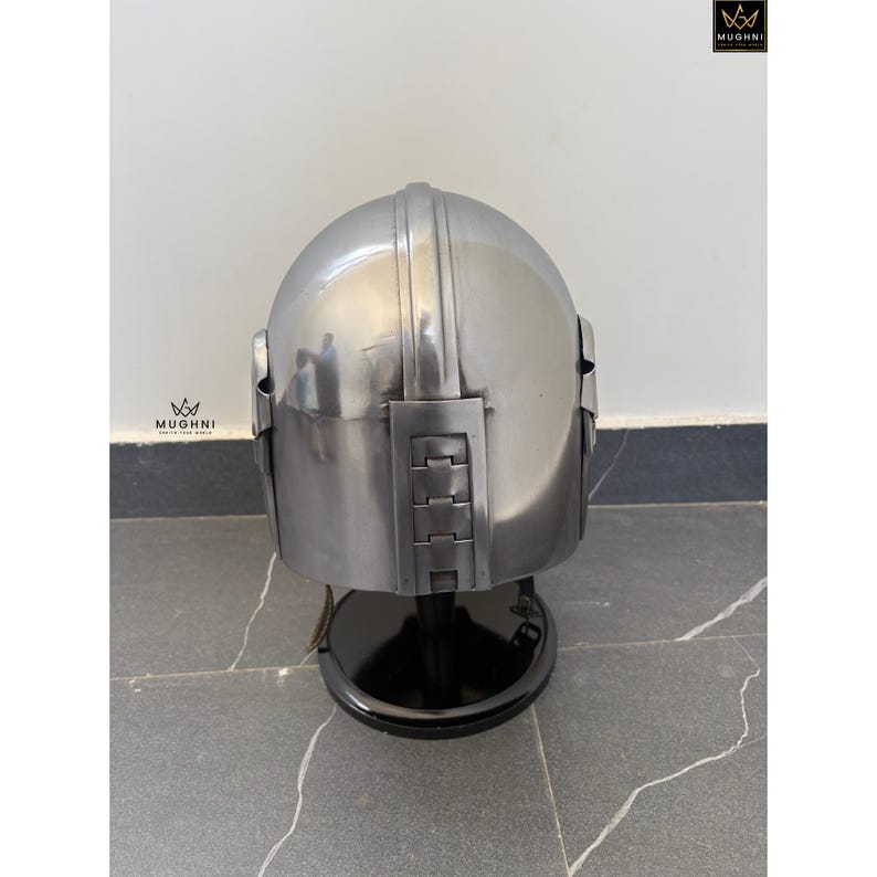 May include: A polished silver helmet with a rounded top and rectangular side and back panels. The helmet is displayed on a black stand. The image includes the logo "MUGHNI" in the upper right and lower left corners.