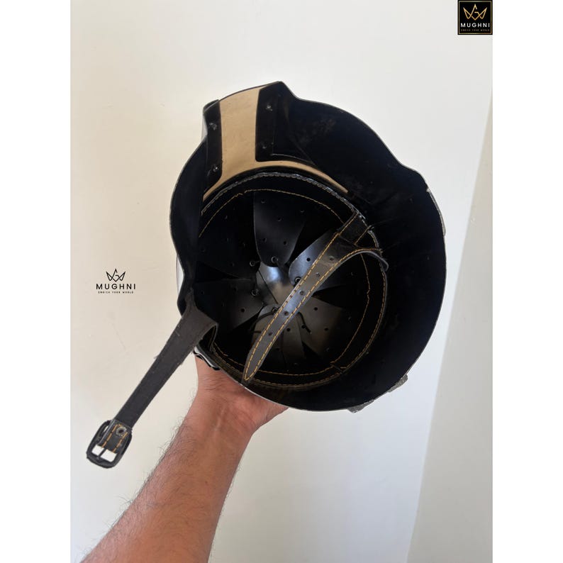 May include: A black and tan medieval helmet with a leather chin strap and interior padding. The helmet has a riveted construction and a polished finish. The interior is black and the exterior is black with tan accents.