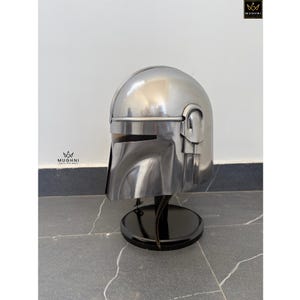 May include: A polished silver helmet with a distinctive visor and ear pieces, displayed on a black stand. The helmet's design is reminiscent of a science fiction aesthetic. The stand is round and has a glossy finish.