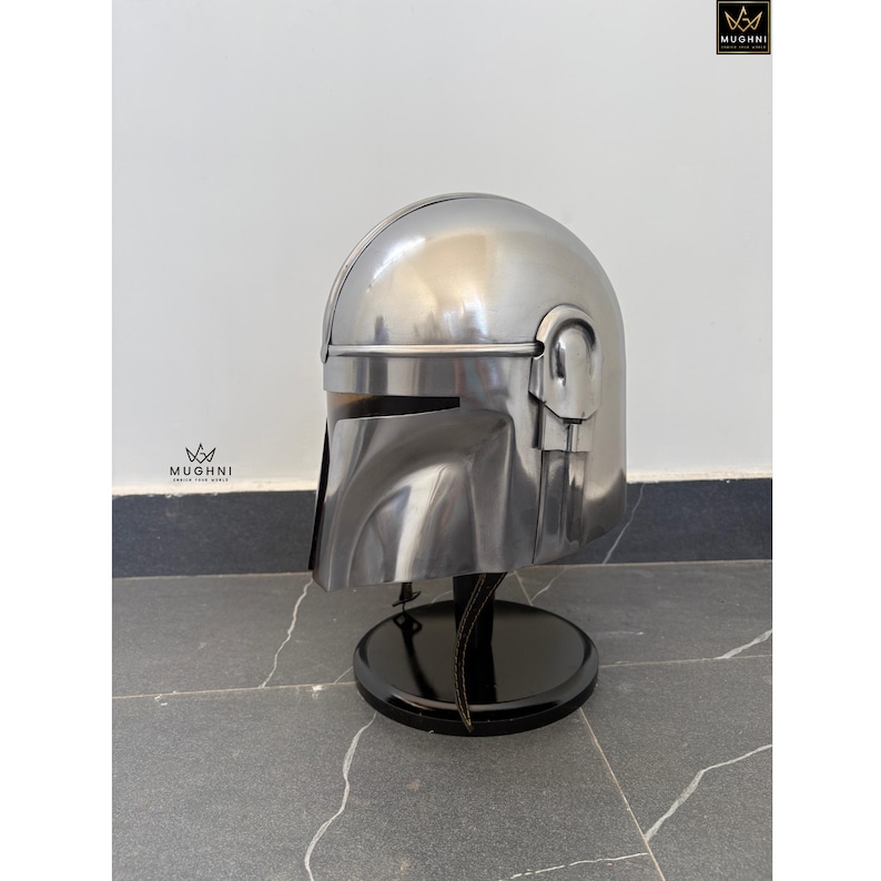 May include: A polished silver helmet with a distinctive visor and ear pieces, displayed on a black stand. The helmet's design is reminiscent of a science fiction aesthetic. The stand is round and has a glossy finish.