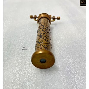 May include: A vintage brass kaleidoscope with an ornate floral design etched on the barrel. The kaleidoscope has a small eyepiece and a set of handles. The brass has a warm, antique finish, and the design is detailed and elegant.