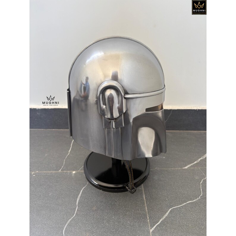 May include: A silver-colored helmet with a rounded top and visor, displayed on a black stand. The helmet has detailed features, including ear pieces and a chin piece. The helmet is made of metal and has a polished finish.