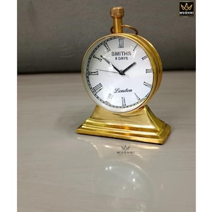 May include: A gold-colored, vintage-style clock with a white face and black Roman numerals. The clock reads "SMITHS 8 DAYS London" and sits on a matching gold base. The clock has a small, gold winding stem at the top.