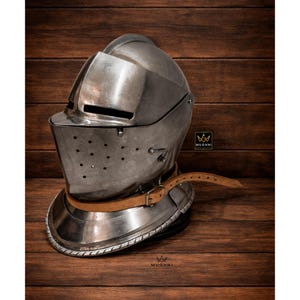 Könnte beinhalten: Ein polierter Ritterhelm aus Stahl mit Visier, Nietdetails und einem Kinnriemen aus Leder. Der Helm liegt auf einer Holzoberfläche und zeigt sein historisches Design und seine Handwerkskunst. Der Helm ist im mittelalterlichen Stil gehalten.