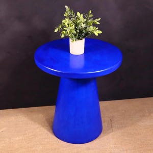 May include: A vibrant blue side table with a round top and a conical base. A small white pot with green leafy plant sits on the table's surface. The table is made of wood and has a smooth finish.
