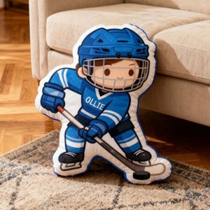 May include: A hockey-themed pillow in the shape of a cartoon hockey player, wearing a blue helmet and uniform. The player is holding a hockey stick and puck. The name "OLLIE" is printed on the jersey. The pillow is on a patterned rug.
