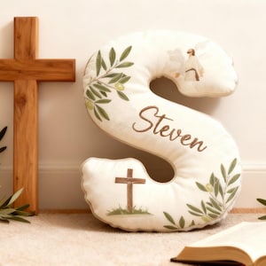 May include: A wooden cross and a cream-colored pillow in the shape of the letter "S" are displayed. The pillow features olive branch and olive illustrations, the name "Steven", and a cross. An open book and olive branches are also present.