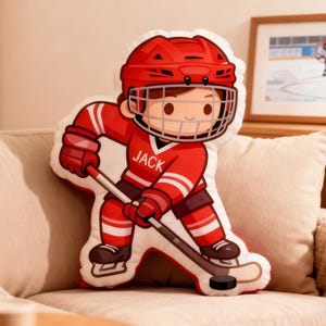 May include: A hockey-themed pillow in the shape of a cartoon hockey player in a red uniform with white stripes. The player is holding a hockey stick and a puck. The name "JACK" is printed on the jersey.