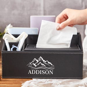 May include: Black faux leather desk organizer with multiple compartments, including a tissue dispenser. The organizer features white stitching and a mountain graphic with the name "ADDISON" in white. A hand is reaching for a tissue.