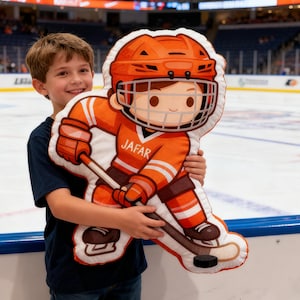 May include: An orange and white hockey player cushion, with the name "JAFAR" on the jersey. The cushion depicts a hockey helmet, gloves, and a hockey stick with a puck. The background shows an ice hockey rink.